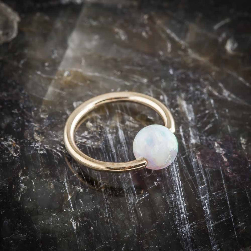 14K yellow gold opal captive bead ring with white opal bead resting on dark mineral stone