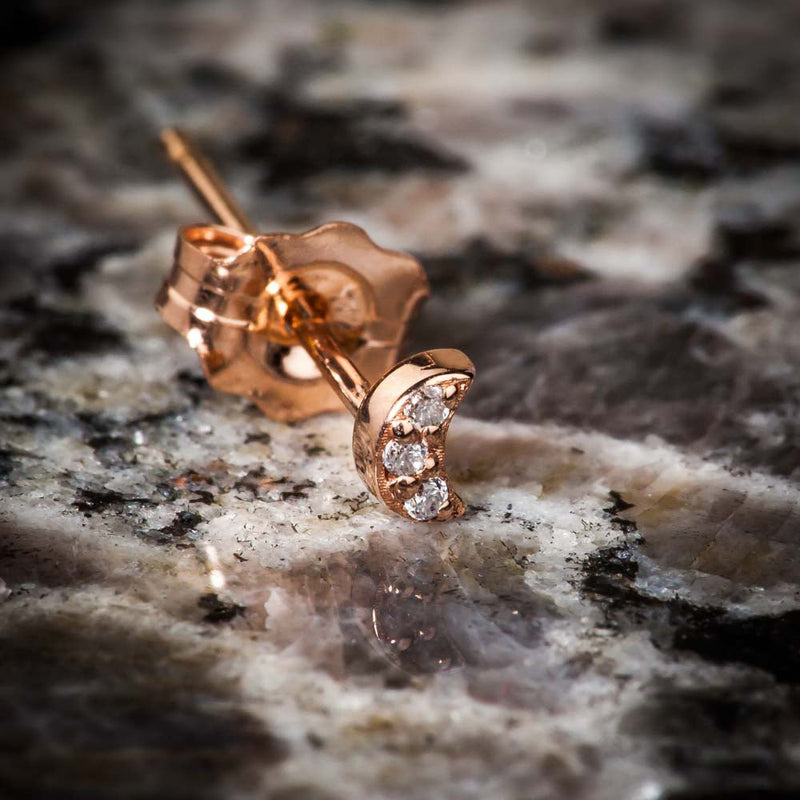 14k rose gold crescent moon diamond stud earring resting on natural stone surface, angled view