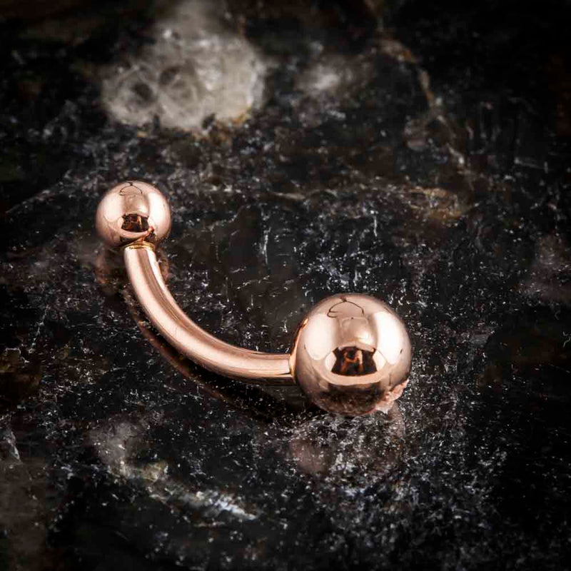 14k rose gold double ball belly ring on dark granite surface, side profile showing curved barbell