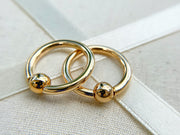 Pair of 14k yellow gold captive bead nipple rings resting on linen textile, angled close-up