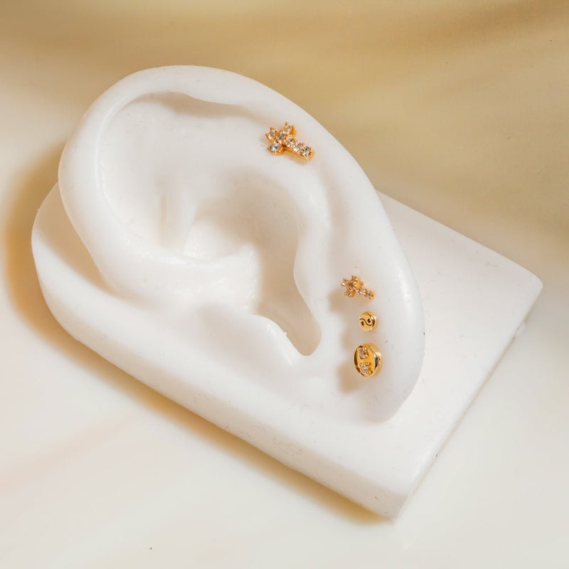 Yellow gold diamond cross cartilage stud shown on white ear form with complementary gold piercing studs