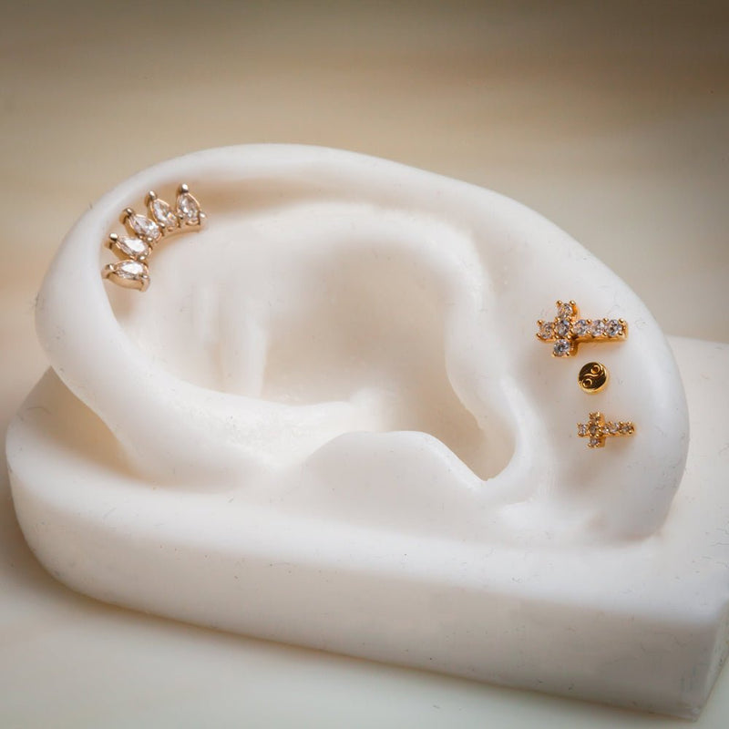 Gold diamond cross labret stud displayed on white ear model beside other gold earrings for size context
