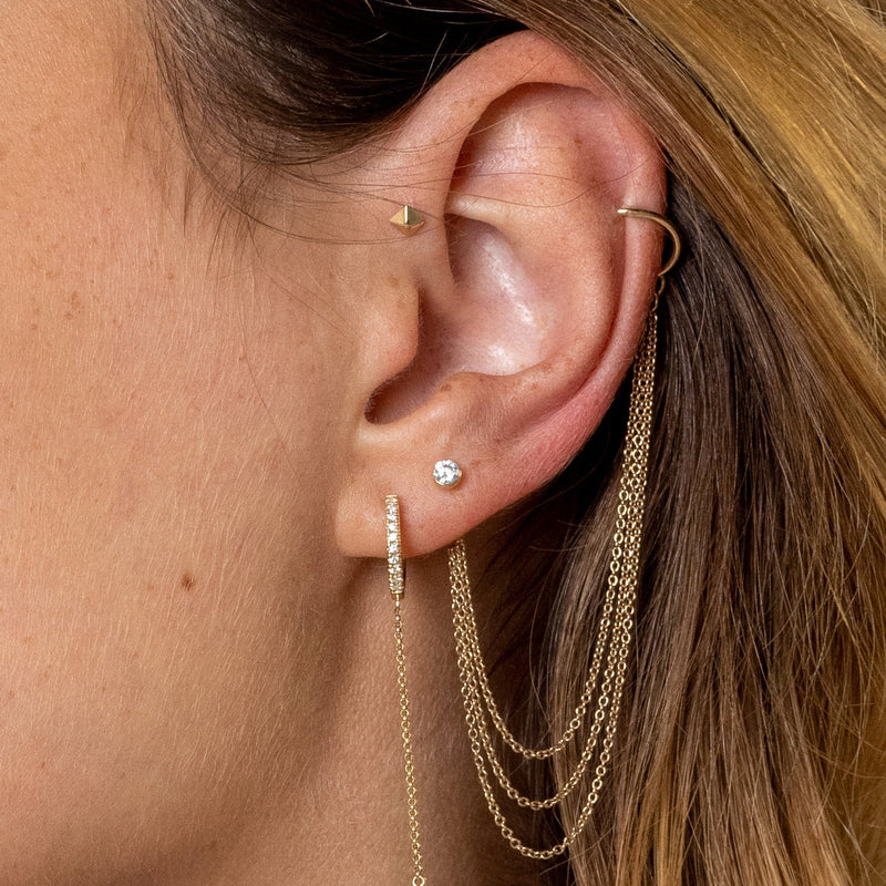 14k yellow gold pyramid stud worn in helix cartilage piercing alongside layered chain earrings on model