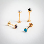 Four 14k gold flat back labrets with pearl, black, amethyst, and turquoise cabochons laid out for size comparison