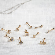 Assorted 14k yellow gold CZ cartilage barbells scattered on white linen, including bezel stud