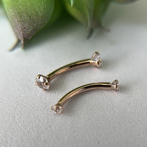 Two 14k rose gold diamond curved barbells resting on a surface with greenery, showing actual size