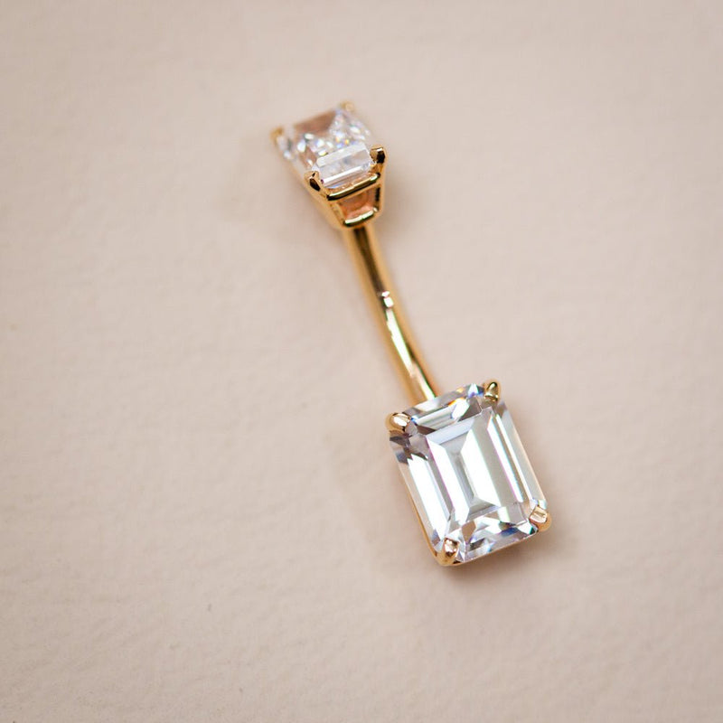 14k yellow gold double emerald-cut clear CZ navel ring laid flat on neutral surface, angled view