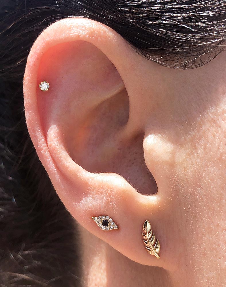 14k yellow gold feather stud worn in second lobe piercing, styled with evil eye and CZ cartilage studs